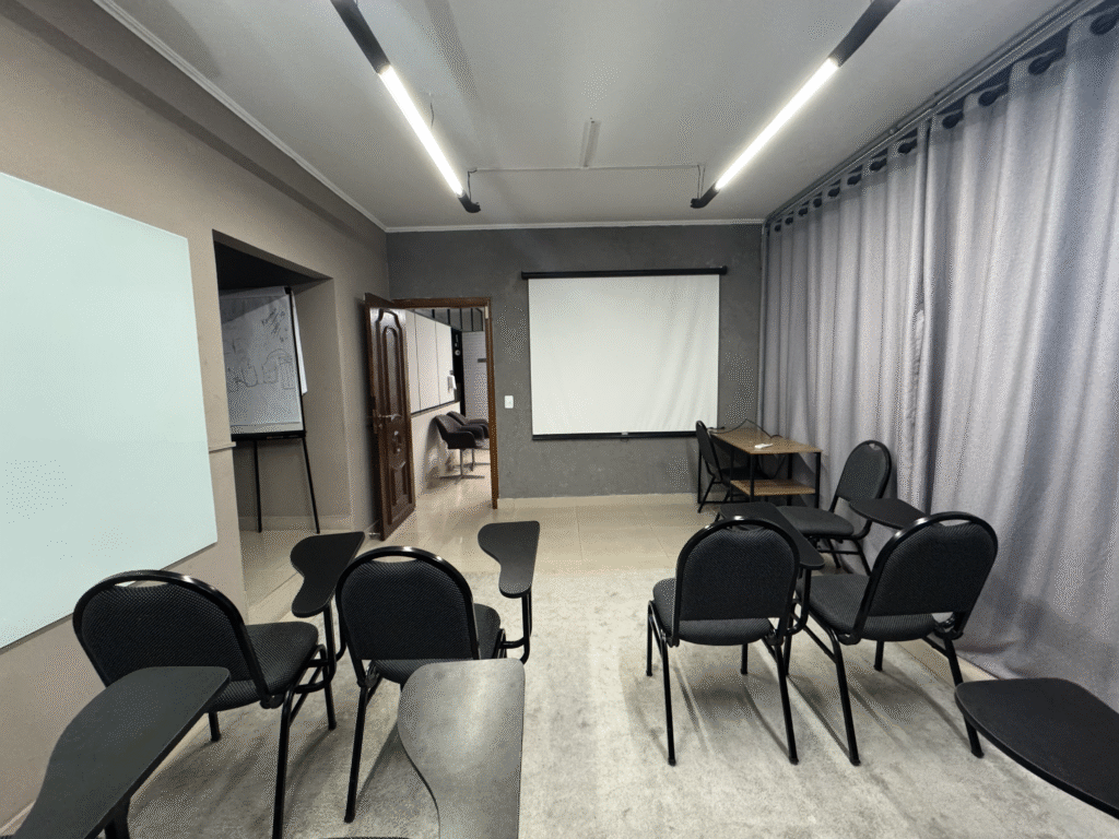 Sala com cadeiras universitarias, quadro branco lateral e tela de projeção